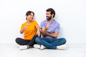 Fototapeta na wymiar Young caucasian couple sitting on the floor isolated on white background giving a thumbs up gesture