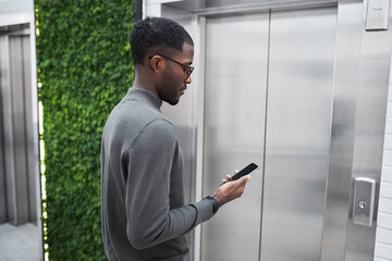 Naklejka na ściany i meble Minimal waist up portrait of young black businessman waiting for elevator in office building and using phone, copy space