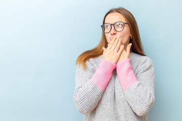 Shocked young woman in eyeglasses covering her mouth with her hands and looking at empty space for your text or object on blue background
