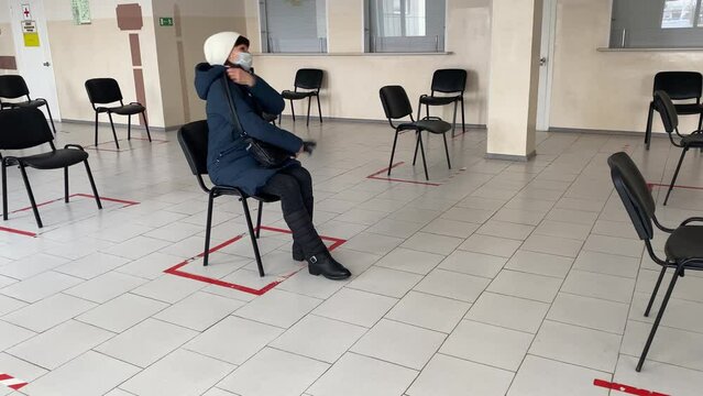 Concept of social distancing in waiting room at station. One masked man is sitting on chair, keeping his distance. Woman leaves leaving an empty hall. New normal life during pandemic.
