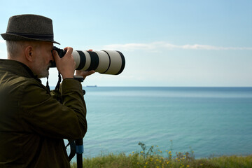 A man photographs a distant marine object with a long-focus lens. The lens is supported on a tripod monopod. Copy space.