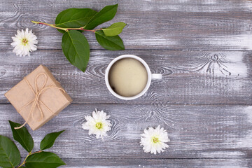 Composition with a cup of coffee, a gift wrapped in kraft paper, a green branch and white chrysanthemum flowers on a gray bleached wooden table. copy space