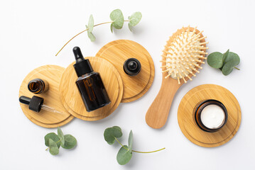 Natural cosmetics concept. Top view photo of wooden hair brush glass bottles and cream jar on wooden stands and eucalyptus on isolated white background
