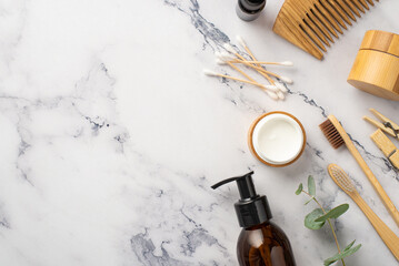 Top view photo of glass bottles wooden hairbrush cream jar toothbrushes cotton buds clothespins and eucalyptus on white marble background with copyspace