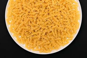 Uncooked Fusilli Pasta Lying on White Plate on Black Background. Raw and Dry Macaroni. Unhealthy and Fat Food - Top View
