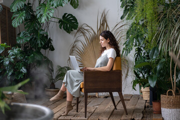 Smiling hispanic female entrepreneur sitting in armchair in greenhouse, working on laptop surrounded by exotic houseplants. Pensive italian woman typing on computer. Cozy workplace, freelance concept © DimaBerlin
