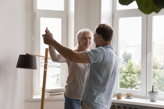 Smiling mature couple in love standing holding hands dance in living room, celebrate life event feeling affection, look happy spend carefree weekend leisure together at home. Relations, date concept