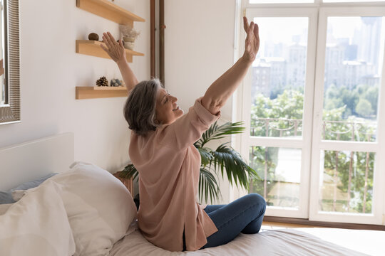 Side view older woman sit on bed in modern light warm bedroom raise hands stretch body muscles feels happy, welcomes new day, enjoy carefree morning, untroubled retired life and comfort living concept