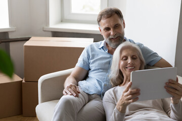 Older couple relax on sofa on move day near boxes with stuff, use tablet buy goods on internet, make order via e-service app, make purchase online, search home renovation new ideas, e-commerce concept