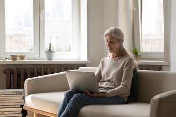 Smiling older hoary woman sit on sofa put wireless computer on laps texting message, chatting remotely through social media networks enjoy distance communication, e-services use modern tech concept