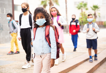 Portrait of positive schoolgirl in mask standing near school, children on background