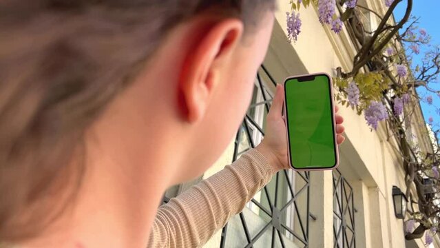Girl looks at the phone lifting it up with chromate blonde natural hair developing green screen flipping her finger against the background of a beige building. High quality 4k footage