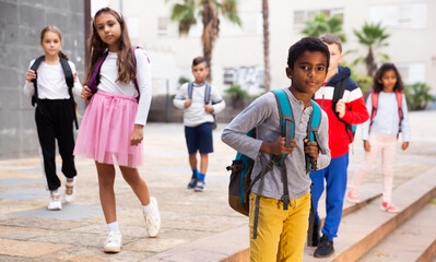 Obraz premium Cute tween african boy with backpack walking with other schoolchildren to school campus after lessons on warm fall day.