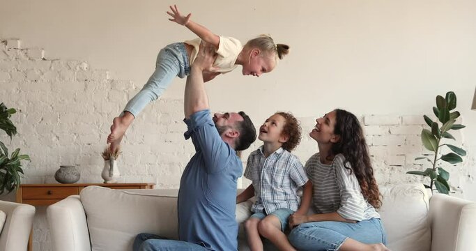 Happy mixed race family play together. Smiling young Latin mom elder son watch strong dad lift younger daughter up in air. Laughing little girl pretend flying bird in funny game with parents brother
