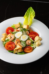 Caesar salad with grilled shrimps, tomato, boiled egg, lettuce, croutons and green sauce. Top view close-up on a brown round plate with fresh vegetables and cutlery by side on a dark background.