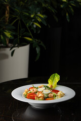 Caesar salad with grilled shrimps, tomato, boiled egg, lettuce, croutons and green sauce. Top view close-up on a brown round plate with fresh vegetables and cutlery by side on a dark background.