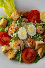 Caesar salad with grilled shrimps, tomato, boiled egg, lettuce, croutons and green sauce. Top view close-up on a brown round plate with fresh vegetables and cutlery by side on a dark background.