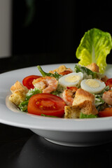 Caesar salad with grilled shrimps, tomato, boiled egg, lettuce, croutons and green sauce. Top view close-up on a brown round plate with fresh vegetables and cutlery by side on a dark background.