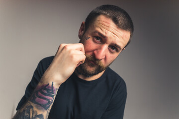 Studio shot of a tattooed guy in his 30s wearing black t-shirt and touching his bushy moustache while looking at camera over gray background. High quality photo