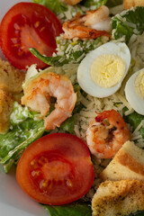 Caesar salad with grilled shrimps, tomato, boiled egg, lettuce, croutons and green sauce. Top view close-up on a brown round plate with fresh vegetables and cutlery by side on a dark background.
