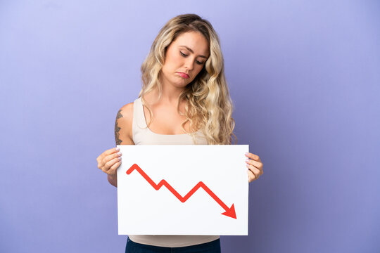 Young Brazilian woman isolated on purple background holding a sign with a decreasing statistics arrow symbol with sad expression