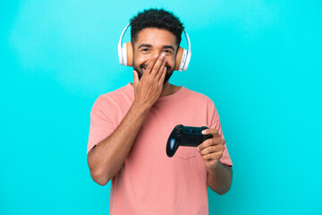 Young brazilian man playing with a video game controller isolated on blue background happy and smiling covering mouth with hand