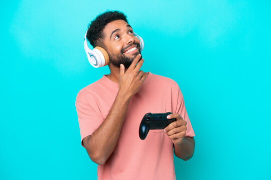 Young brazilian man playing with a video game controller isolated on blue background looking up while smiling