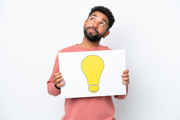 Young Brazilian man isolated on white background holding a placard with bulb icon and looking up