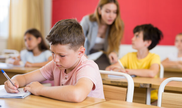 Portrait of concentrated tween boy studying in classroom, listening to schoolteacher and writing in notebook