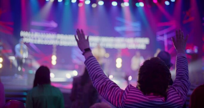 Unrecognizable Christians Worship and Praise God with Raised Hands on Music Concert, Youth Festival or Conference. Believers People Sing Songs on Big Live Stage Event. 4K backside view handheld shot