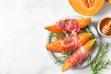 Melon cantaloupe slices with prosciutto ham, rosemary and honey in a plate on white background. top view