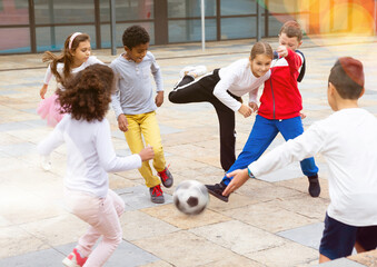 Happy tween girls and boys of different nationalities playing football in schoolyard during break...