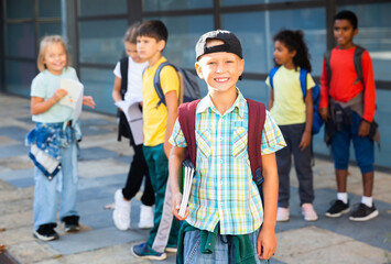 Obraz premium Cute cheerful tween boy looking at camera while standing in school campus with notebooks in hands on background with schoolmates.