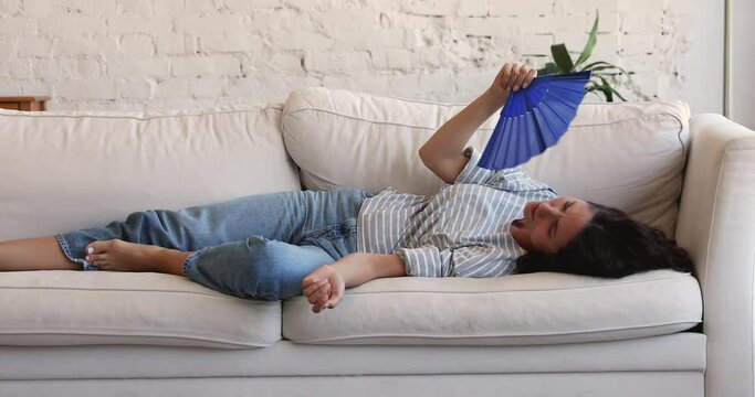 Sad upset young Hispanic lady lie on sofa at home make cool breeze using hand fan suffer of extreme heat think of buying modern air conditioning system. Exhausted woman complain on hot dry air indoors