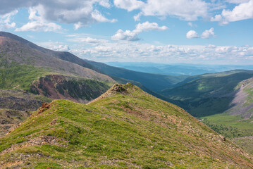 Obraz premium Scenic view from mountain ridge to green forest valley among mountain ranges and hills on horizon at changeable weather. Green landscape with sunlit mountain vastness under cumulus clouds in blue sky.
