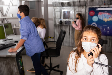 Fototapeta na wymiar Tween girl in protective face mask talking on telephone while her friends trying to solve conundrums in closed space of quest room bunker