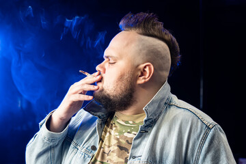 Man with mohawk on head smokes a white cigarette at night on a black background in the smoke....