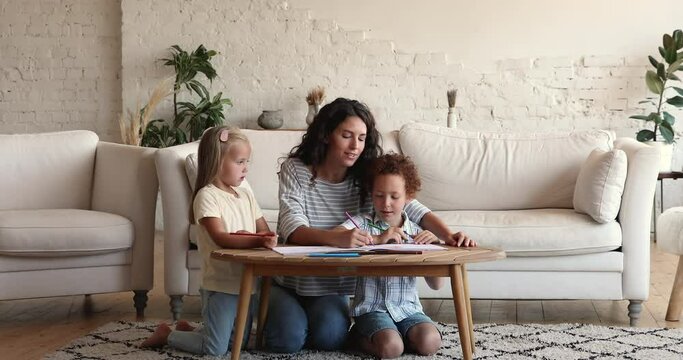 Young Latin female nanny play with two little children diverse siblings at living room teach to draw color sketch using pencils. Happy mom assist kids brother sister in creating picture at paper album