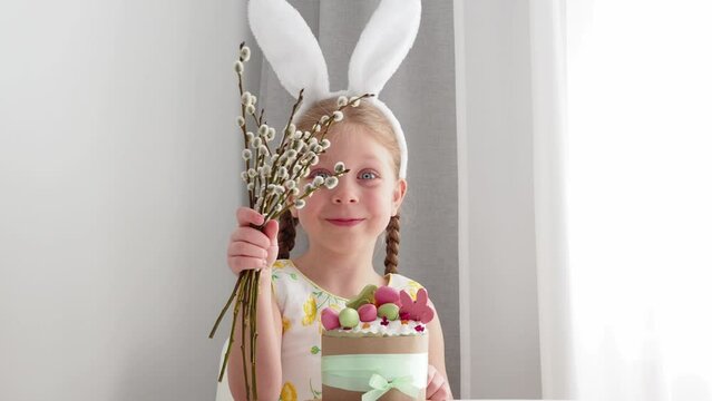 A beautiful, cute, smart girl in an Easter bunny costume smiles and holds a willow bouquet in her hand. symbol of the holiday of light easter - painted eggs, easter cake, willow, easter rabbit.