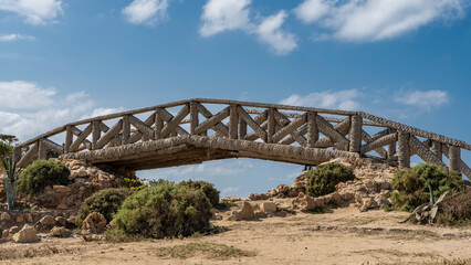 Obraz premium The decorative arch bridge is made of palm trunks. Lattice railings against a background of blue sky and clouds. Boulders and green vegetation are visible on the soil. Montazah Park. Alexandria. Egypt