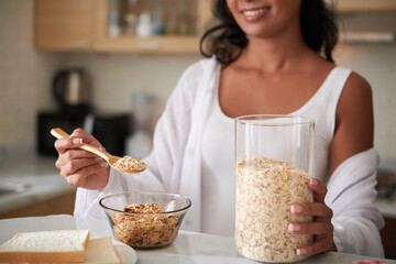 Young woman adding spoon of oat flakes in bowl when making tasty and healthy breakfast