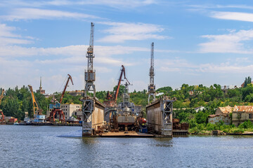Fototapeta na wymiar Repair platform of Mykolayiv Shipyard - view from the Pedestrian bridge across the Inhul river in Mykolaiv, Ukraine. Workshops and harbor cranes against the backdrop of a green shore on a summer day