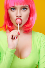 Hipster woman in pink wig with beautiful breasts eat lick lollipop on bright yellow background