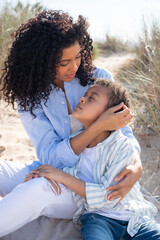 Fototapeta premium Caring African American mother and child on beach. Mother and son in casual clothes sitting on blanket, hugging, eyes closed. Family, relaxation, nature concept