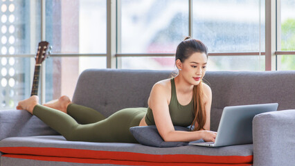 Millennial Asian young healthy slim strong sexy female sporty model in casual crop top sportswear laying down smiling on sofa in living room learning yoga workout class via laptop computer.