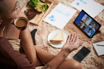 Young entrepreneur drinking cup of morning coffee when talking to colleagues in online meeting