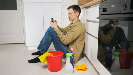 Young husband takes break after cleaning kitchen sitting on floor near bucket and cleanser. Tired housekeeper checks mail on mobile phone at home