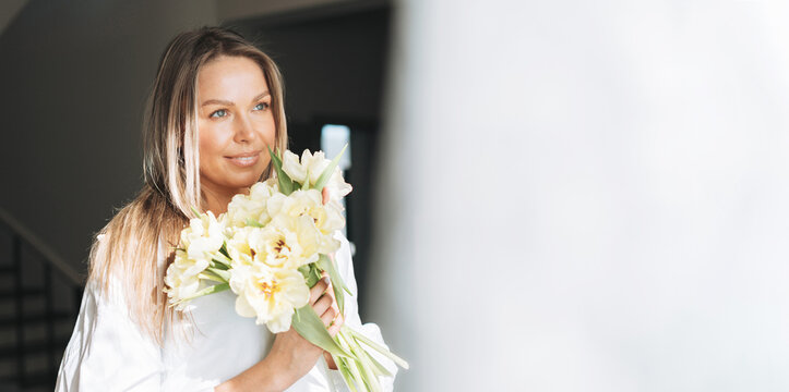 Young smiling woman with blonde long hair in white shirt with bouquet of yellow flowers in hands near window in bright interior, banner