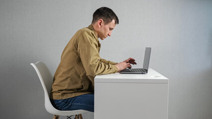 Young man fidgets on chair working on laptop against grey wall. Employee in mustard jacket feels uncomfortable sitting at table in office side view