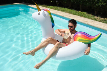 Young man with sunglasses drinks a refreshment on a unicorn inflatable ring.